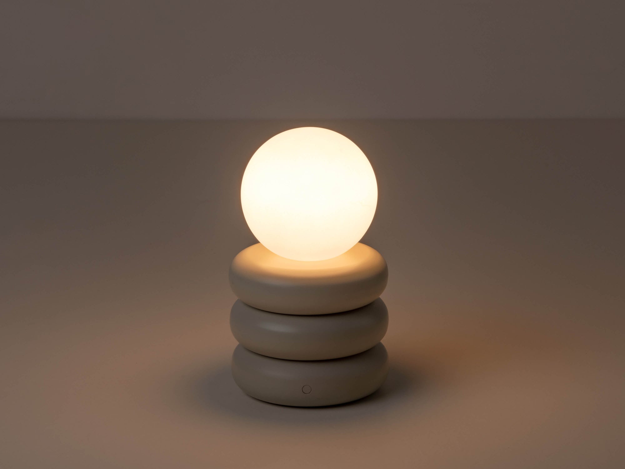 A cordless lamp consisting of three sand-coloured metal tubular discs supports an opal glass shade. The opal glass shade contains an integrated LED bulb that can be recharged. The lamp is portable and wireless. The light is on in a darkened room.