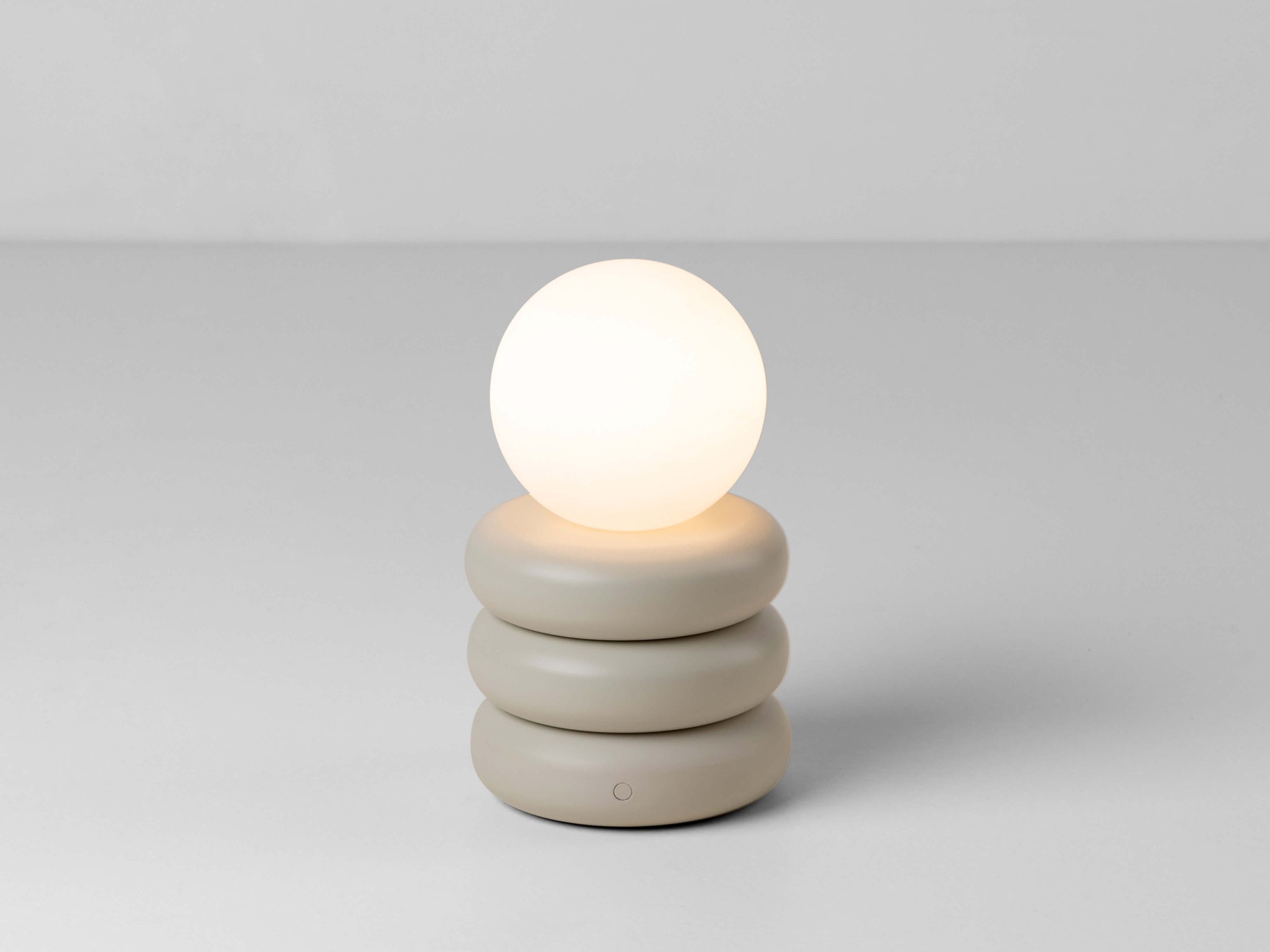 A cordless lamp consisting of three sand-coloured metal tubular discs supports an opal glass shade. The opal glass shade contains an integrated LED bulb that can be recharged. The lamp is portable and wireless. The light is on.