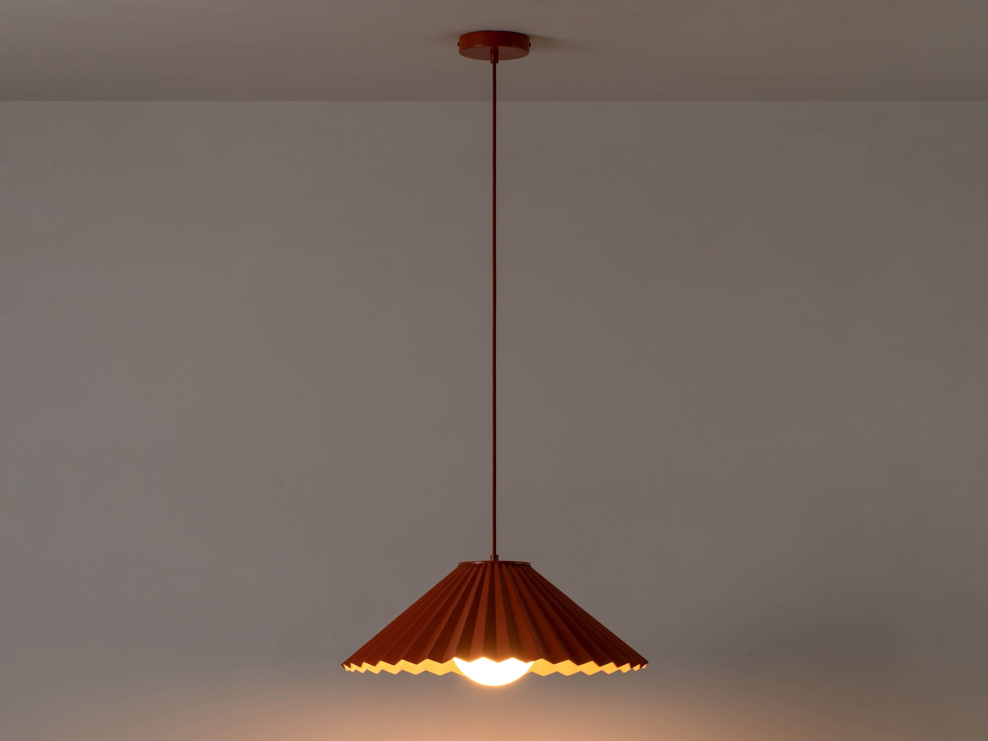 A large, fluted, pleated metal shade is suspended from a ceiling pendant. It is painted burnt orange on the outside, and pale pink on the inside. Light is on and in a darkened room.