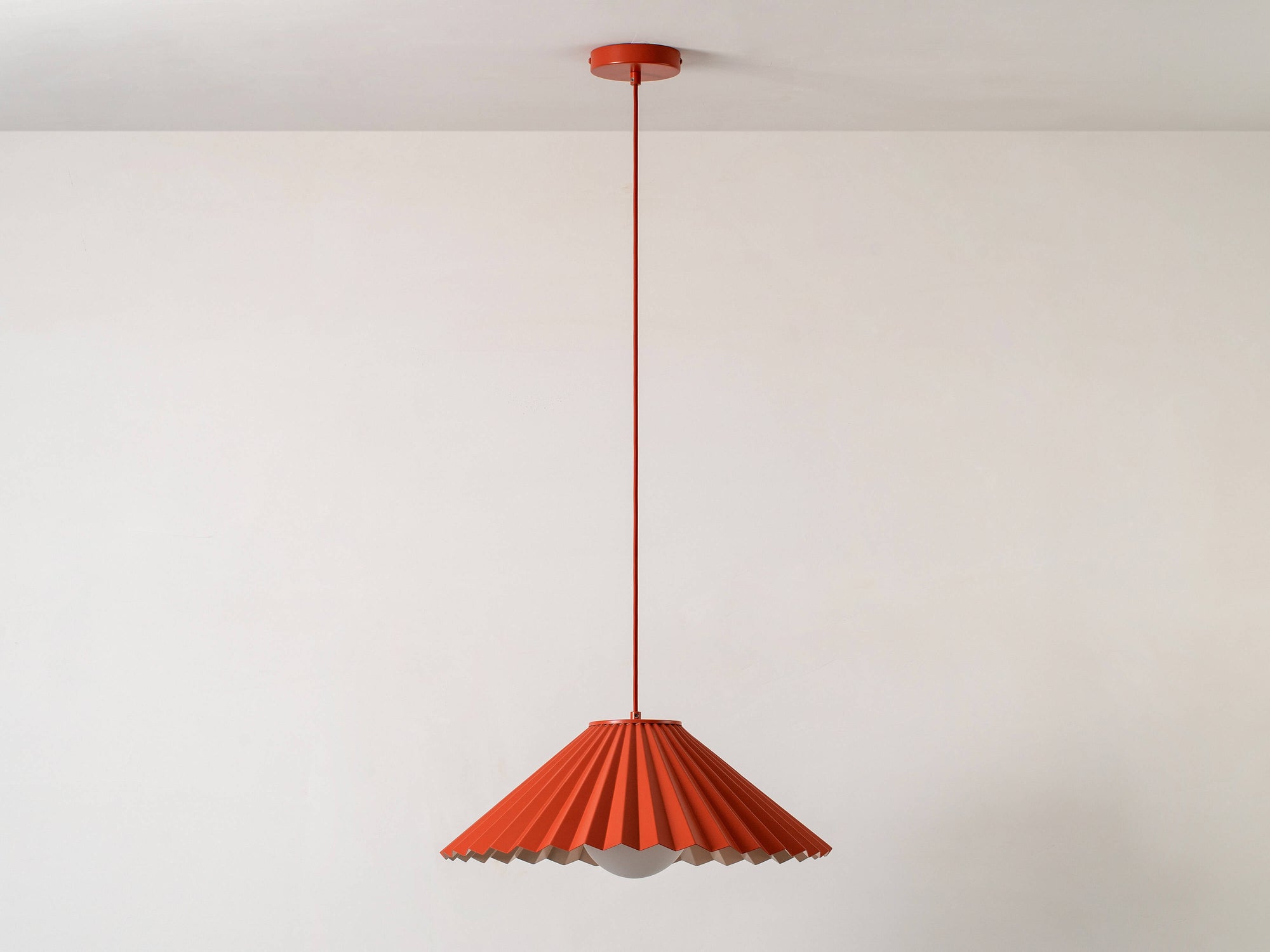 A large, fluted, pleated metal shade is suspended from a ceiling pendant. It is painted burnt orange on the outside, and pale pink on the inside. Light is off.