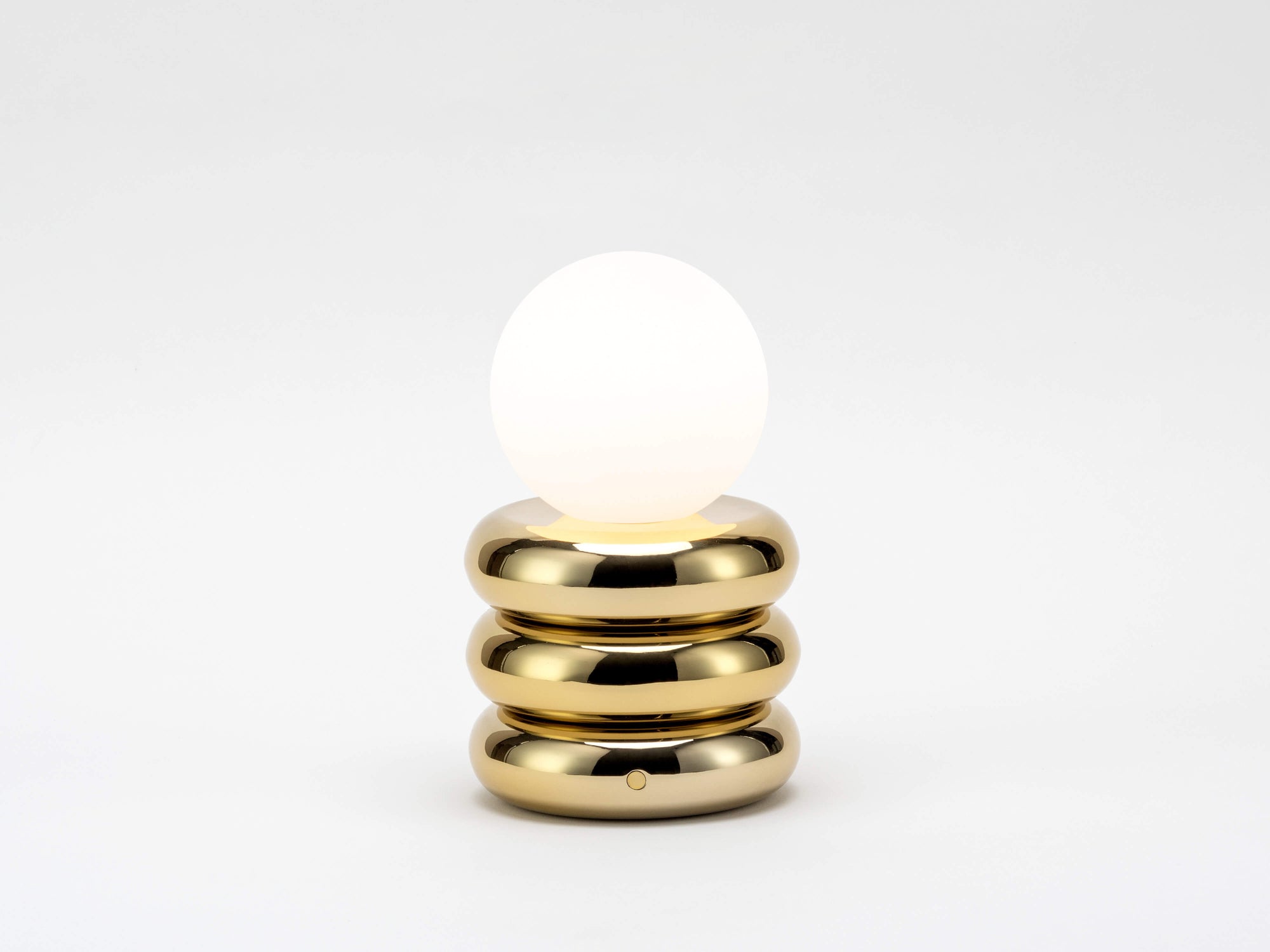 A cordless lamp consisting of three brass metal tubular discs supports an opal glass shade. The opal glass shade contains an integrated LED bulb that can be recharged. The lamp is portable and wireless. The light is on.