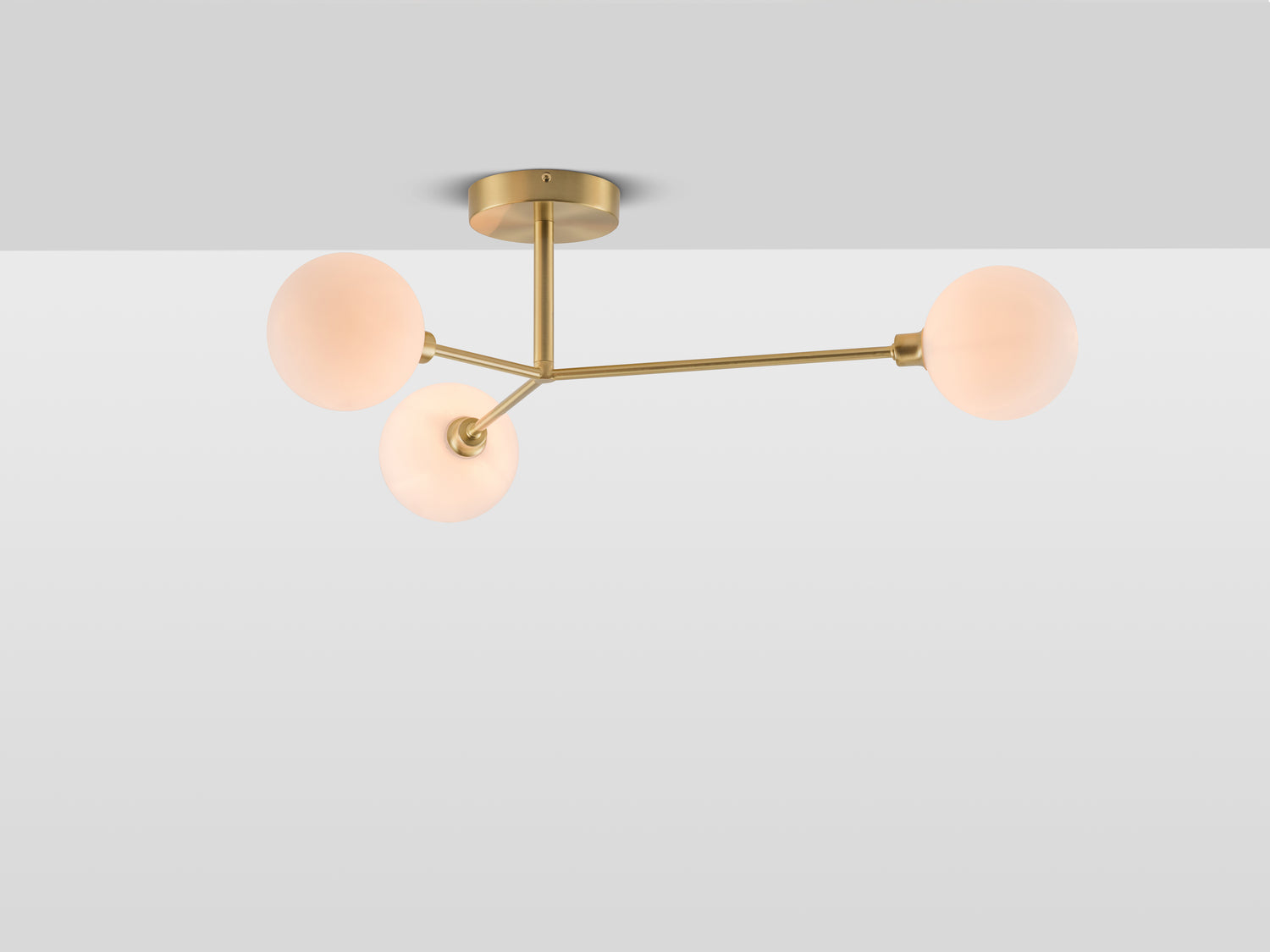 A flush-fit brass ceiling light with three brass metal stems of varying lengths, each suspending a large white opal glass shade. Lights on.